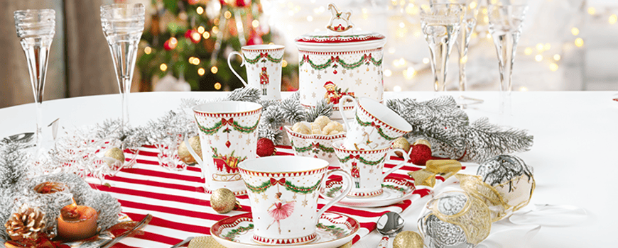Christmas table set against a blurred background of a Christmas tree, showcasing the cups, mugs, and a sweets jar from the Magic Christmas Collection (Easy Life). All items feature the ballerina and toy soldier theme and are arranged on a red and white striped table runner, flanked by pine garlands with silver and gold ornaments. Tall, elegant champagne flutes are visible in the background, highlighting the festive composition.